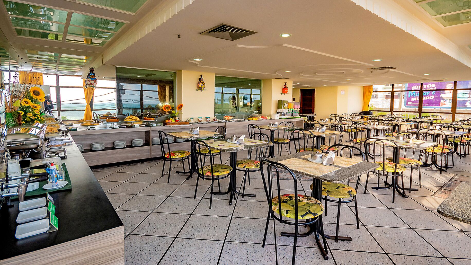 A spacious dining area with multiple tables and chairs, a buffet counter with various food items, and large windows providing a view of the outdoors.