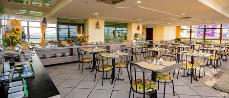 A spacious dining area with multiple tables and chairs, a buffet counter with various food items, and large windows providing a view of the outdoors.