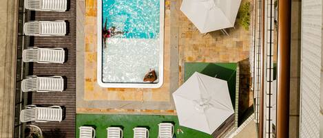 An aerial view of a hotel pool area with a rectangular pool, white lounge chairs, and umbrellas.