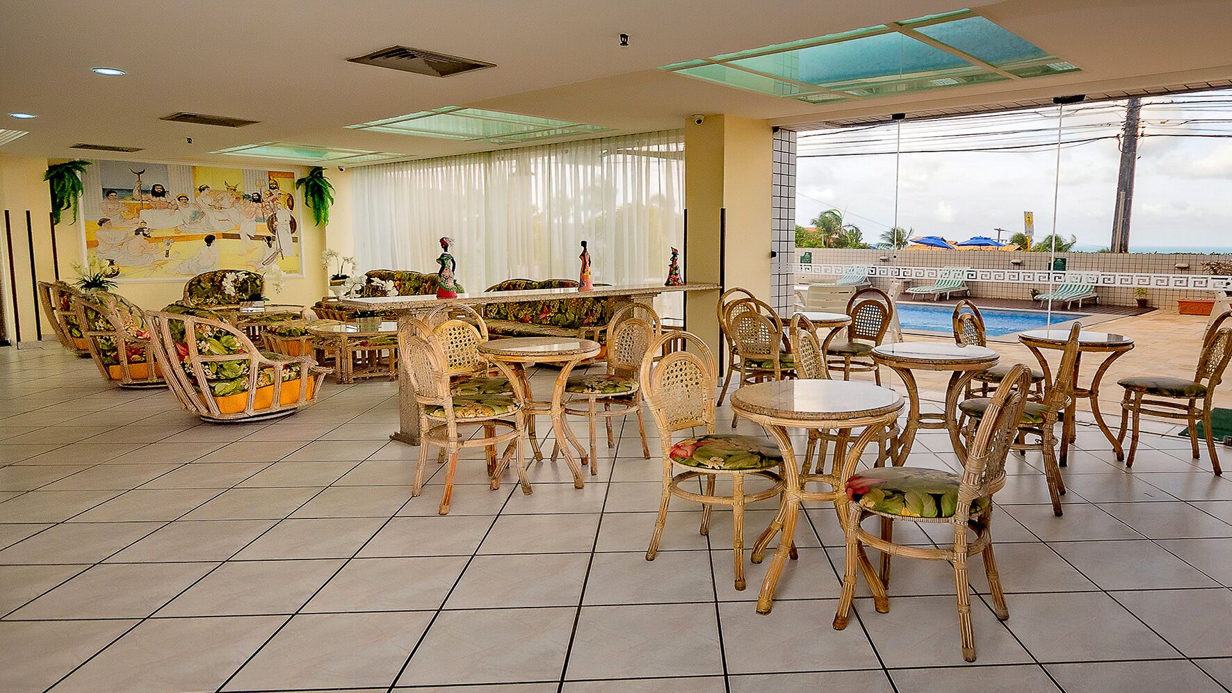 A spacious indoor area with multiple wicker tables and chairs, a large window offering a view of the outdoors, and a tiled floor.