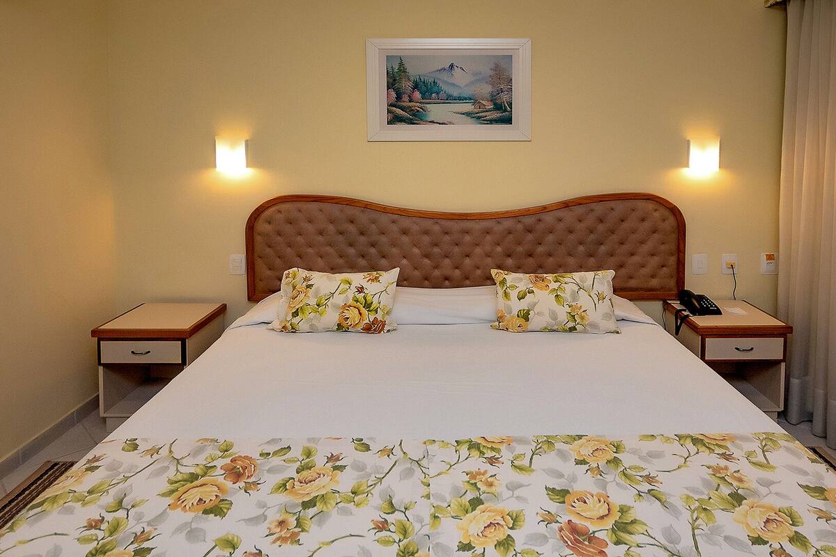 A hotel room with a large bed, floral bedspread, two nightstands, a telephone, and a painting on the wall.