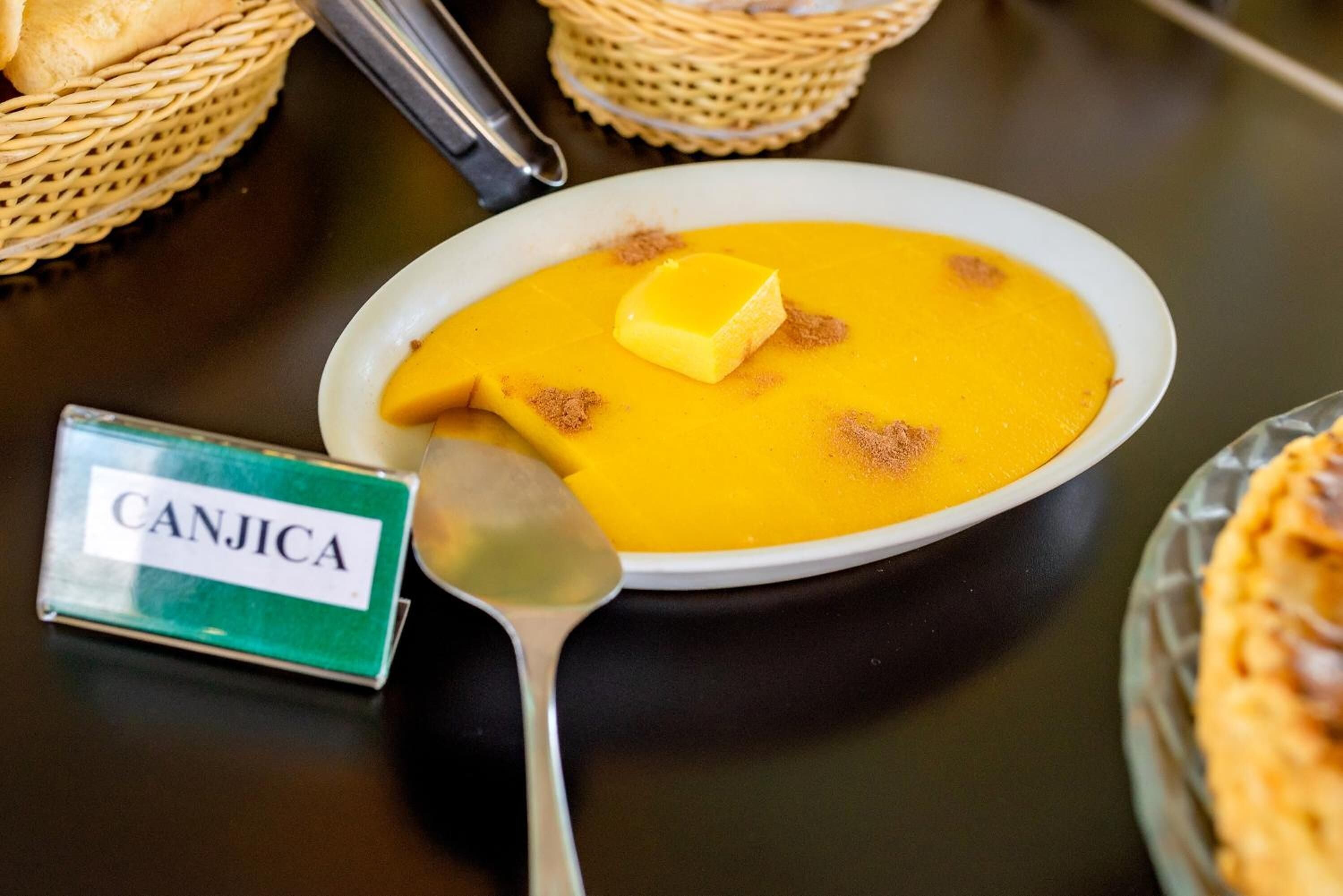 Uma tigela de sopa com o rótulo 'CANJICA', com uma colher e um pedaço de manteiga por cima.