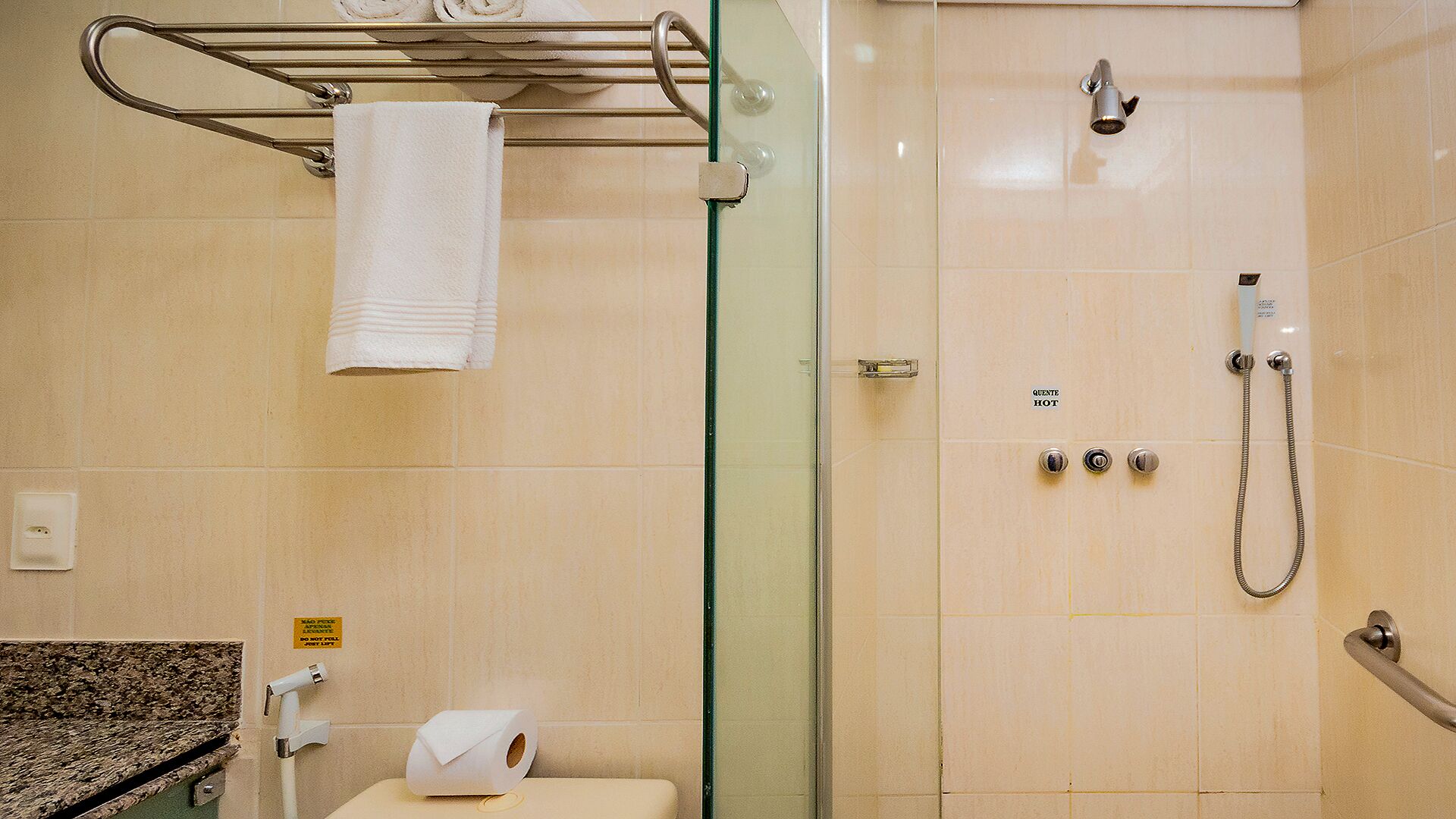 A bathroom with a glass shower enclosure, a towel rack with a rolled towel, a toilet with a paper towel dispenser, and a sink with a faucet.