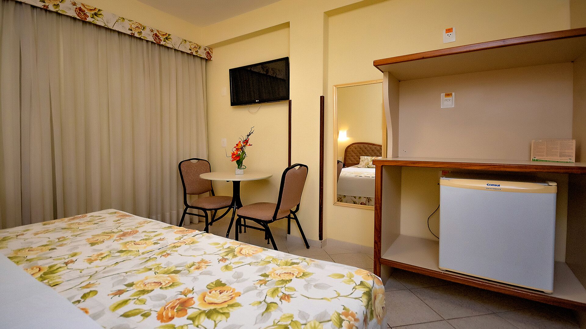 Ein Hotelzimmer mit einem Bett, einem Schreibtisch mit Computer, einem Sessel, einem kleinen Tisch mit Blumenvase und einem an der Wand montierten Fernseher.