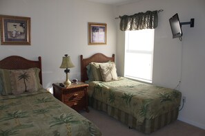 A bedroom with two single beds, a nightstand with a lamp, a wall-mounted TV, and a window with blinds.