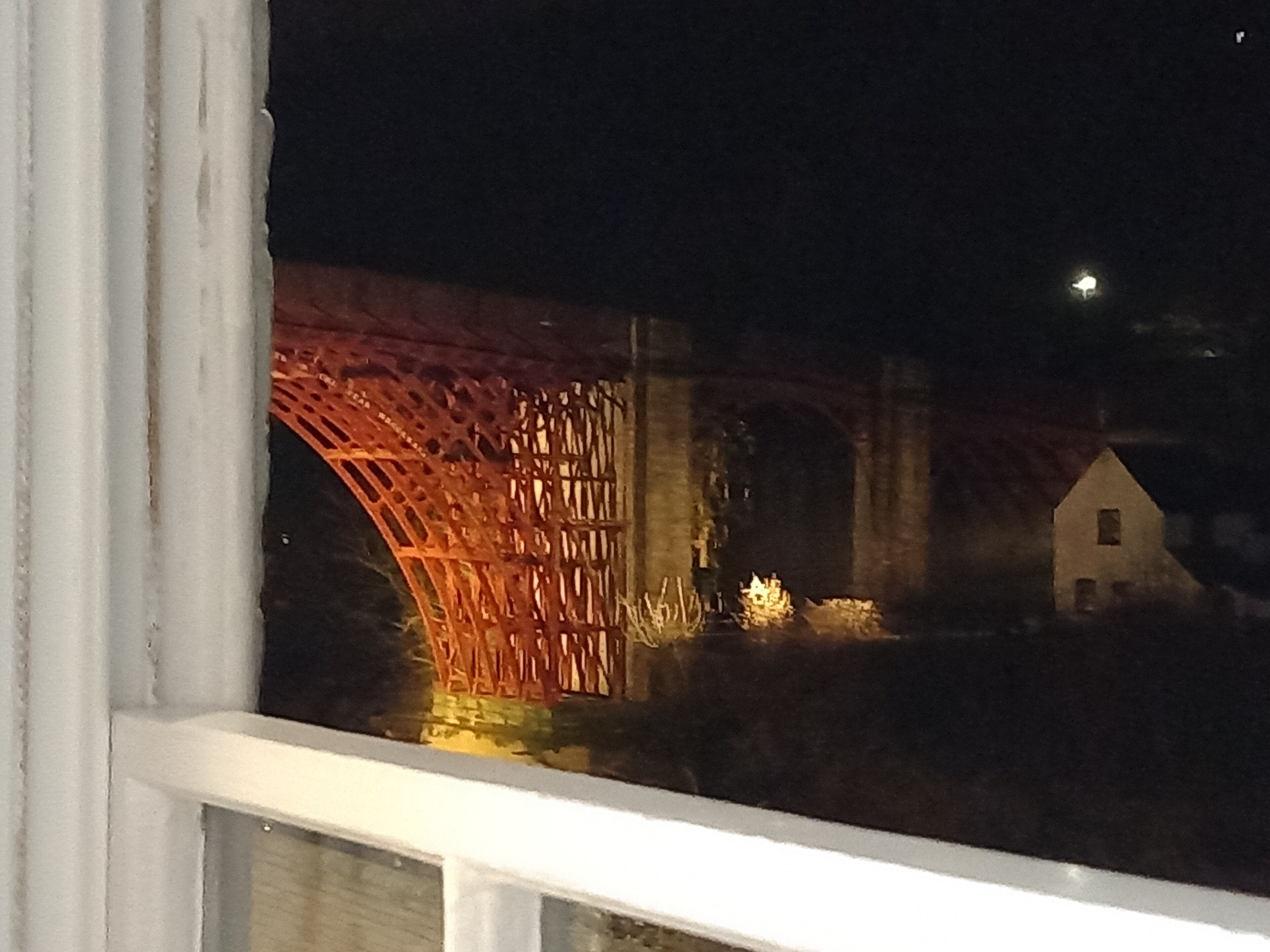Blick auf eine Brücke bei Nacht, mit einer beleuchteten Struktur und einem kleinen Haus im Hintergrund.