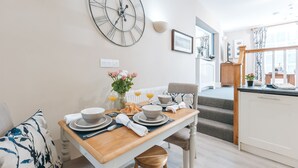 A dining area with a wooden table set for breakfast, a large wall clock, and a view of the kitchen and living area. - Ironbridge View Townhouse (Telford)