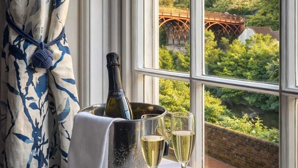 A table with a bottle of champagne, a bucket, and two glasses, set against a window with a view of a bridge and greenery. - Ironbridge View Townhouse (Telford)