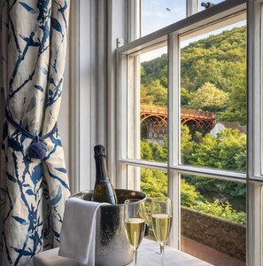 A table with a bottle of champagne, a bucket, and two glasses, set against a window with a view of a bridge and greenery. - Ironbridge View Townhouse (Telford)