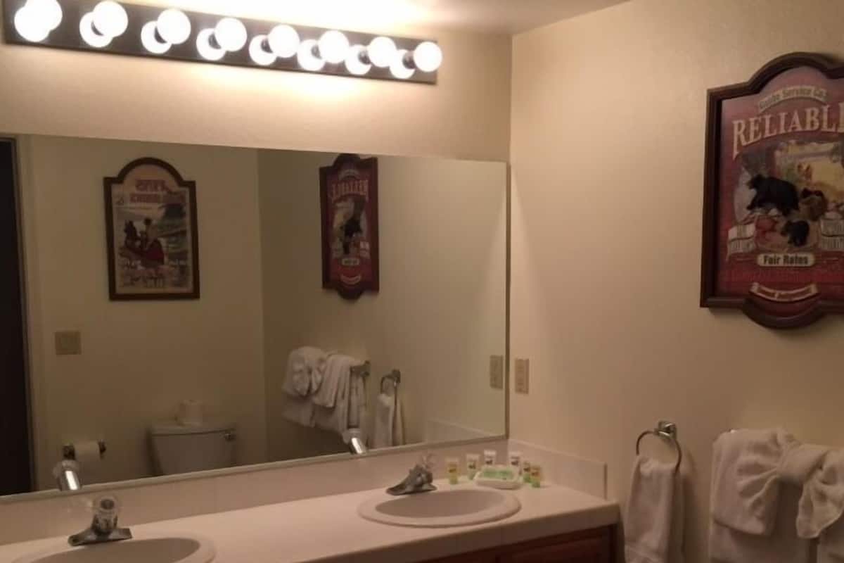 A bathroom with a large mirror, two sinks, and a vanity with drawers. There are towels hanging on the wall and a poster above the mirror.