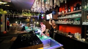 Uma área de bar com balcão, prateleiras com copos e garrafas, e luminárias cilíndricas penduradas.
