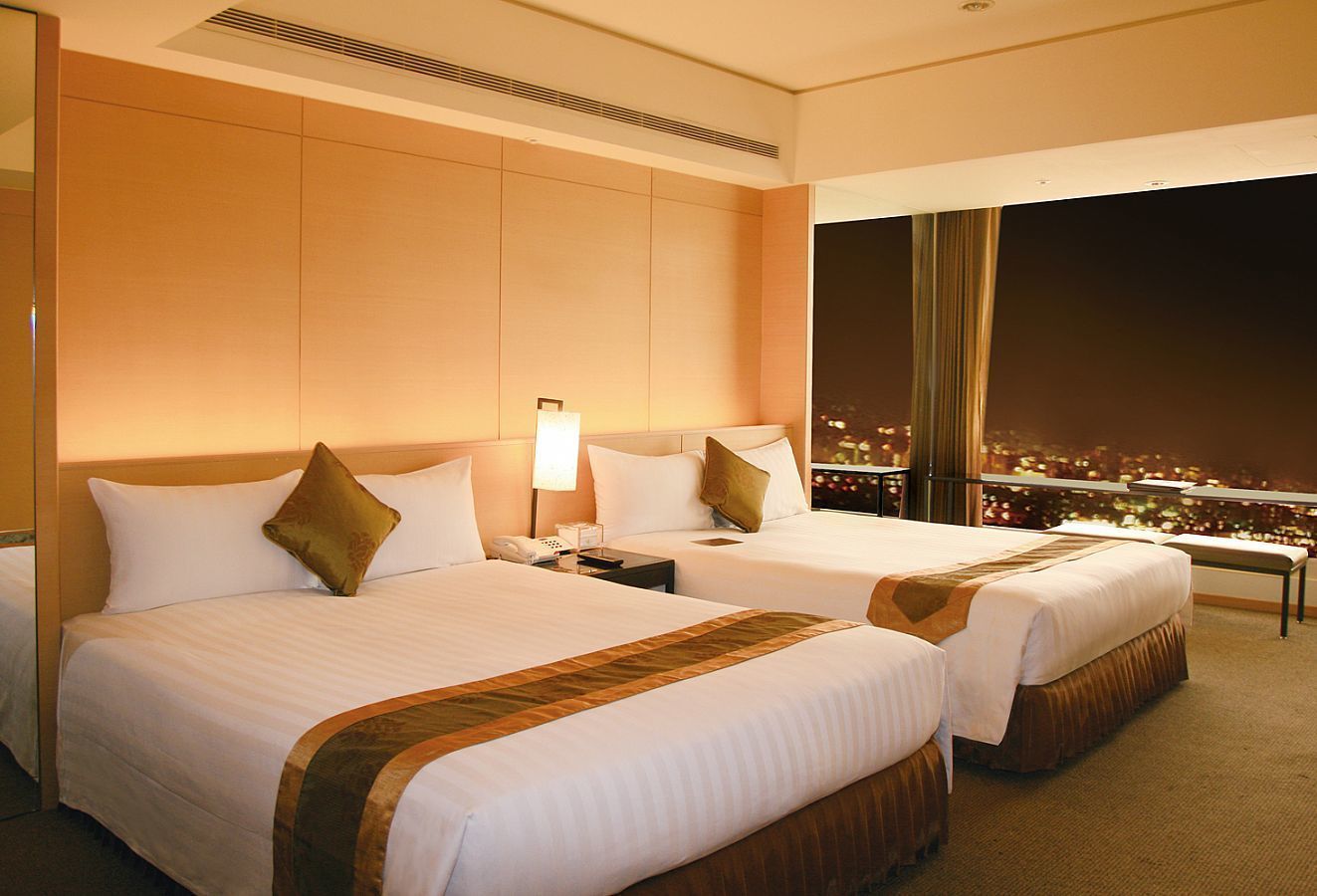 Habitación de hotel con dos camas, una mesita de noche, una lámpara y vista a la ciudad de noche.