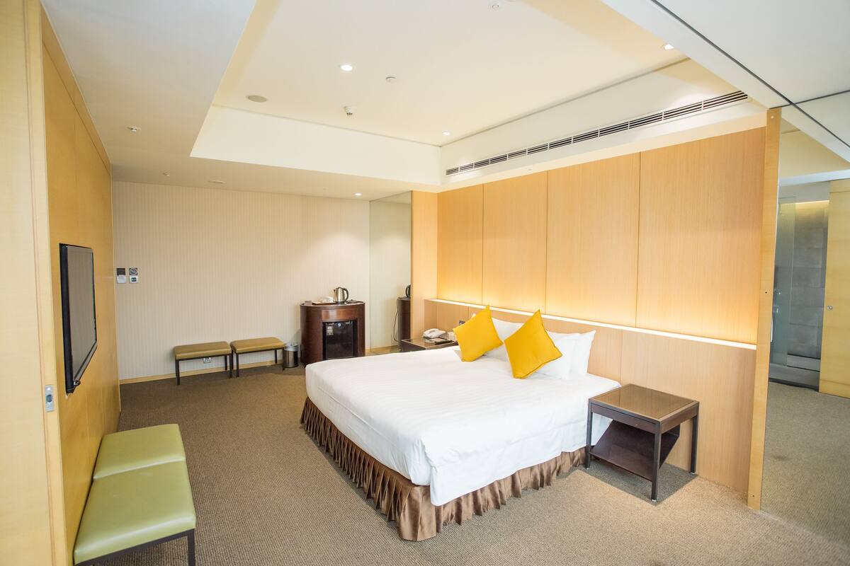 A hotel room with a large bed, a bedside table, a bench, a small table with a phone, and a wall-mounted TV.