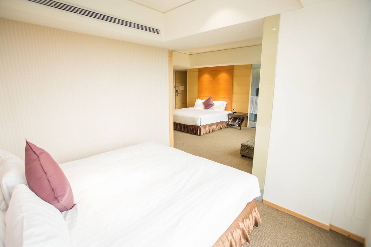 A hotel room with a large bed, a bedside table, and a view into another room.