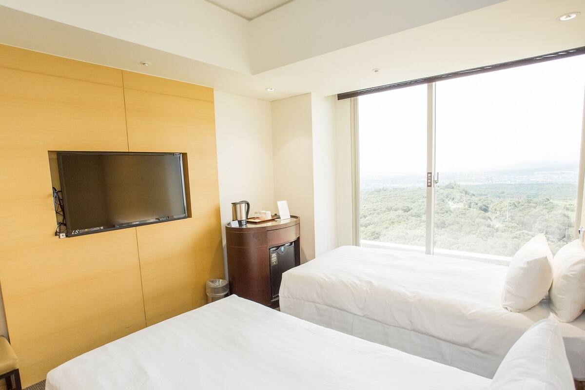 A hotel room with two beds, a TV mounted on the wall, a small table with a kettle, and a large window offering a view of the landscape.