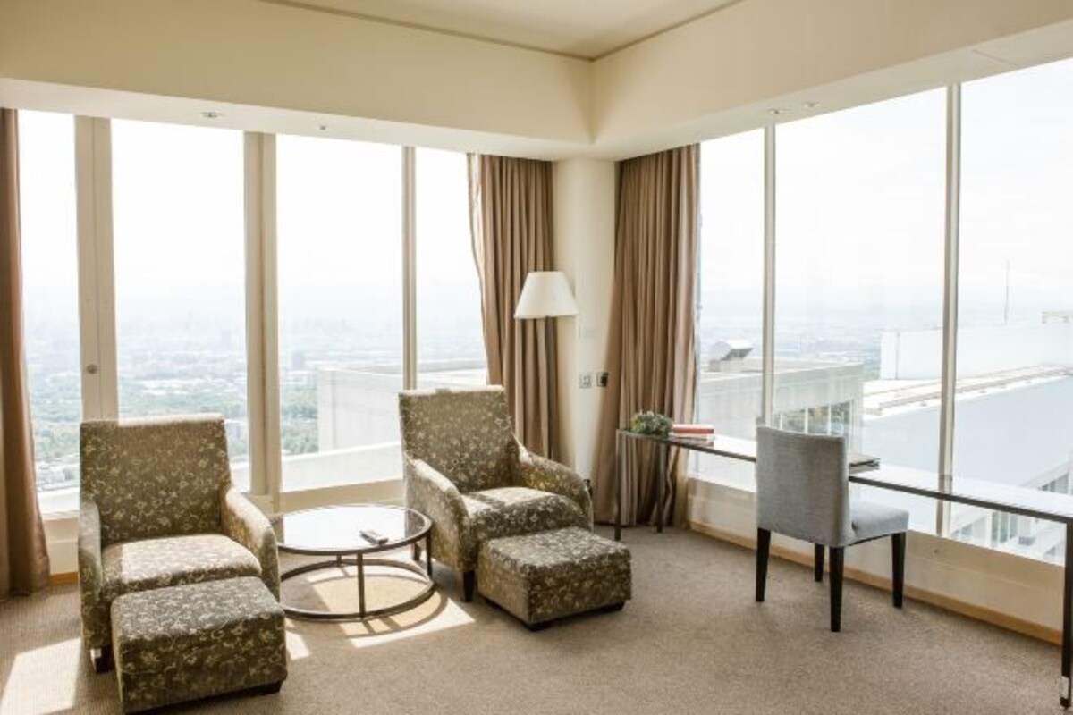 A high-rise hotel room with large windows, a sofa, a chair, a small table, and a lamp.
