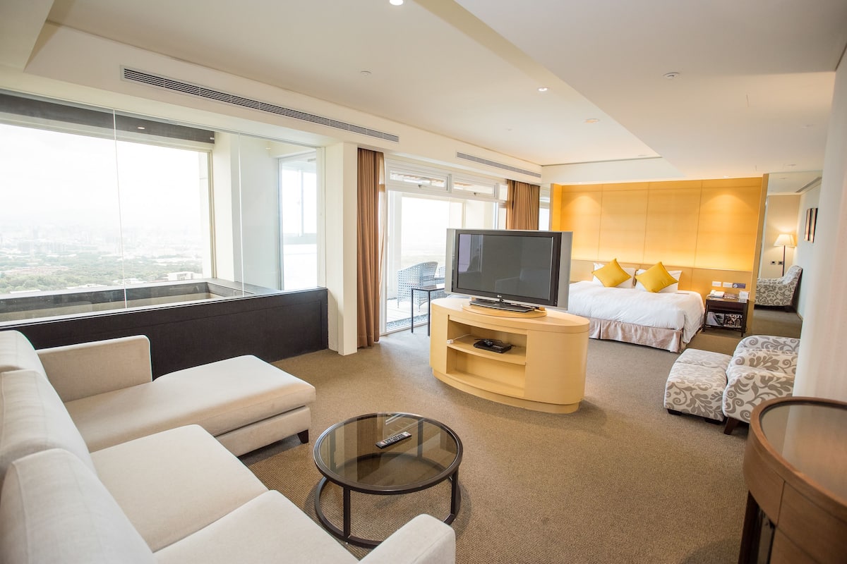 A modern hotel room with a large window, a flat-screen TV, a sofa, a bed with pillows, and a small table.