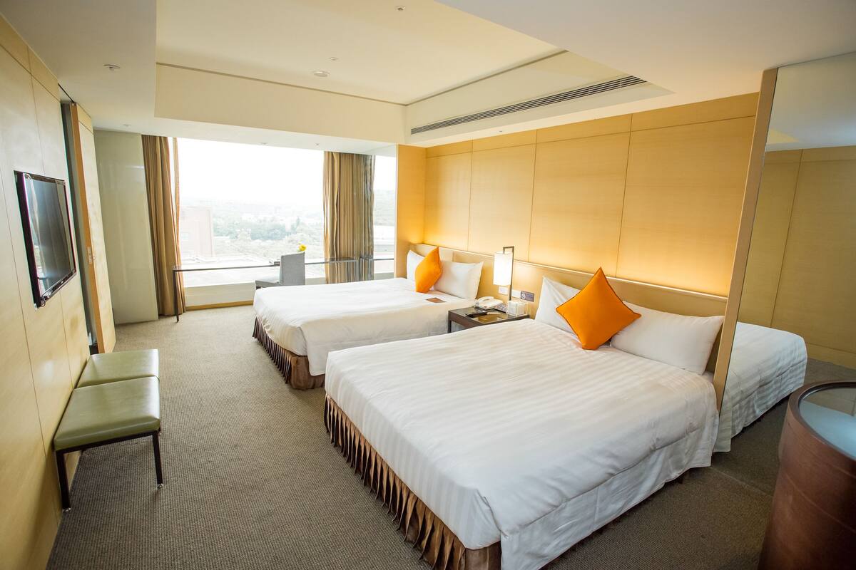 A hotel room with two beds, a small table, a chair, a TV, and a large window with curtains.