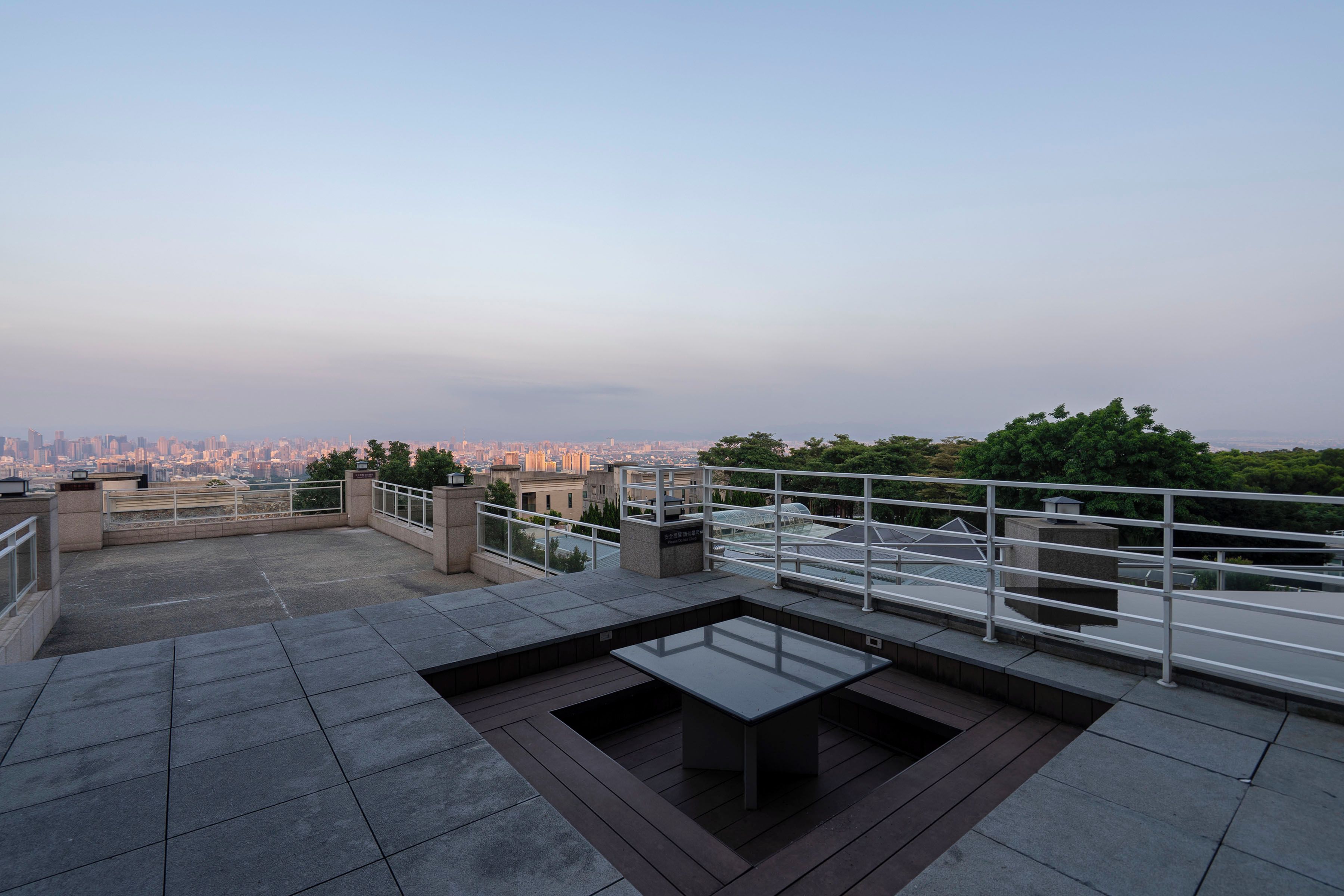 A rooftop terrace with a view of a cityscape, a glass table, and a metal railing.