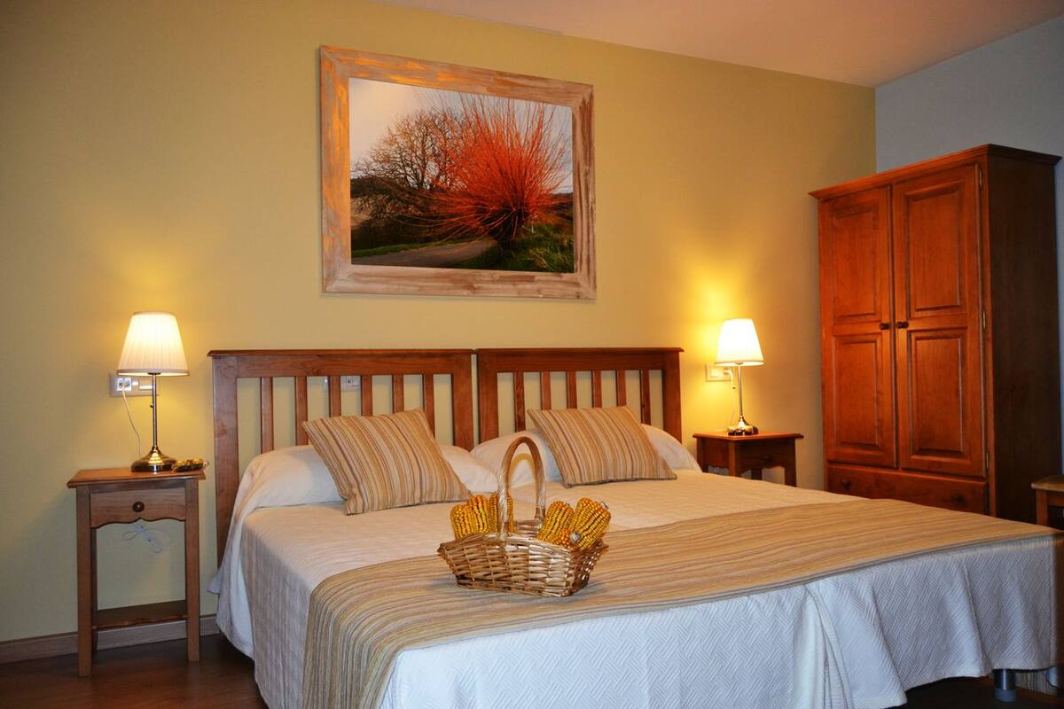 A neatly made bed with a striped bedspread, two bedside lamps, a wooden headboard, a wooden wardrobe, and a framed painting above the bed.
