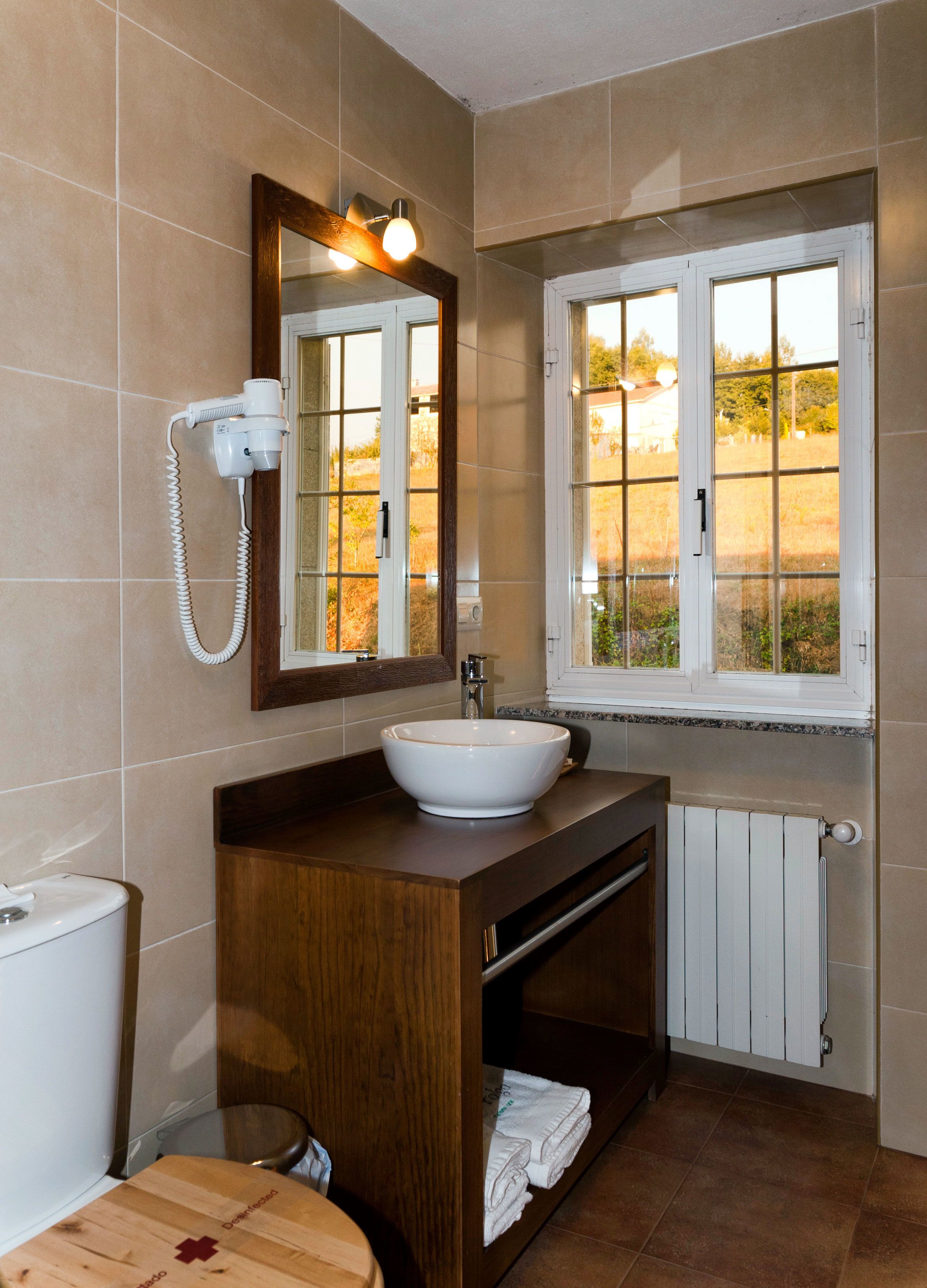 A bathroom with a white sink, wooden vanity, a mirror with lights, a window, and a toilet.