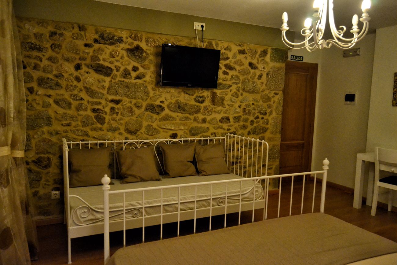 Une chambre avec un mur en pierre, un lit avec un cadre en métal blanc, un téléviseur fixé au-dessus du lit et un lustre.