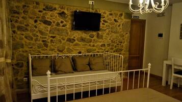 Une chambre avec un mur en pierre, un lit avec un cadre en métal blanc, un téléviseur fixé au-dessus du lit et un lustre.