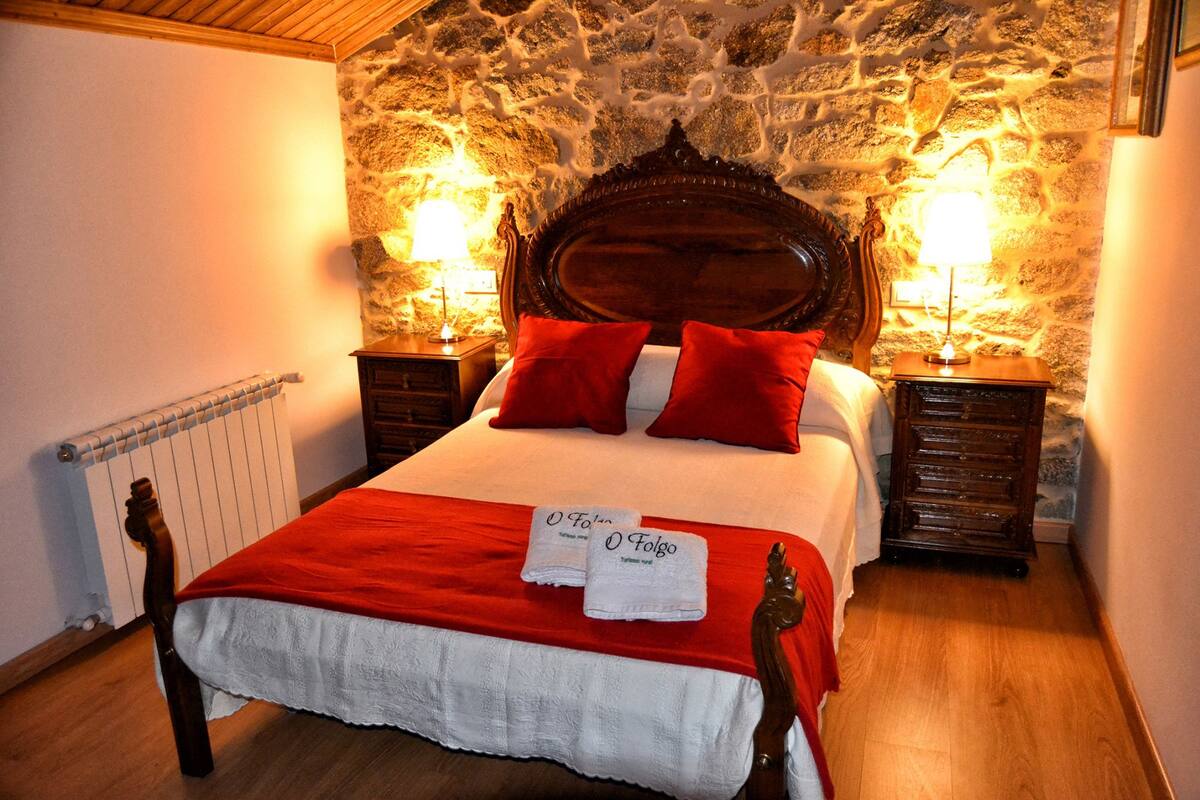 A bed with a red blanket, two red pillows, and two towels with "O Folgo" text.