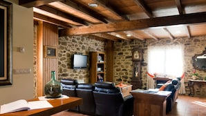 A cozy living room with stone walls, a wooden ceiling, a television, a fireplace, and a dining area. - A Casa do Folgo (A Baña)