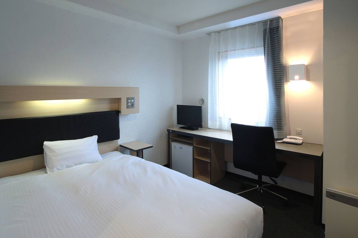A hotel room with a bed, desk, chair, and a small refrigerator.