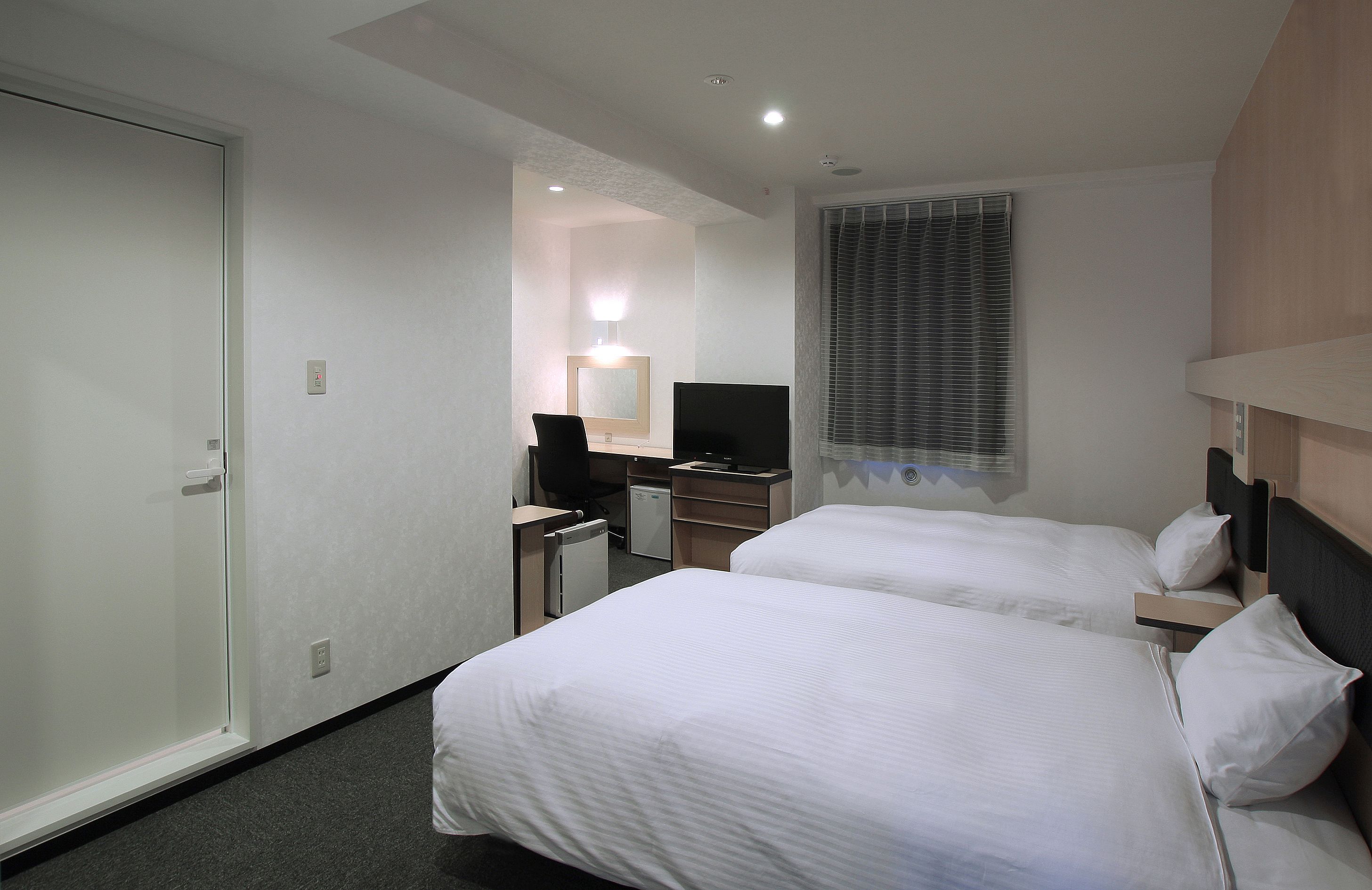 A hotel room with two beds, a desk with a computer, a TV, and a window with blinds.
