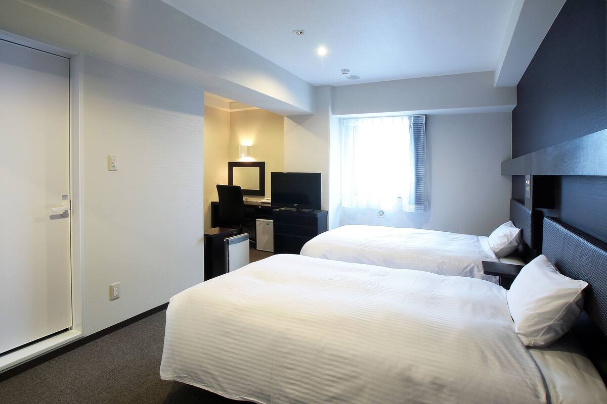 A hotel room with two beds, a desk with a computer, a TV, and a window with blinds.