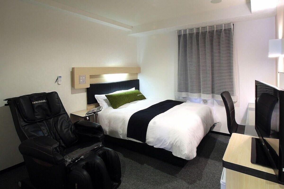 Ein Hotelzimmer mit Bett, Massagesessel, Schreibtisch und einem Fenster mit Jalousien.