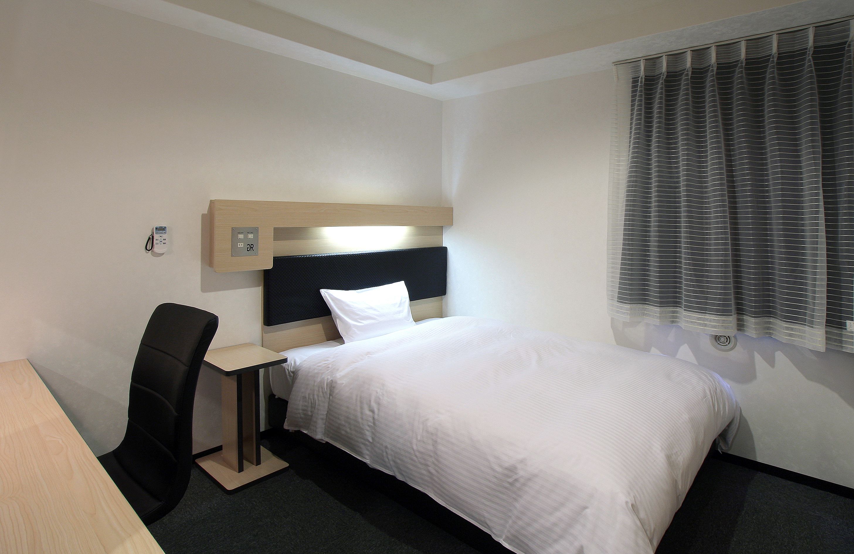 A hotel room with a bed, a desk, a chair, a bedside table, and a window with curtains.