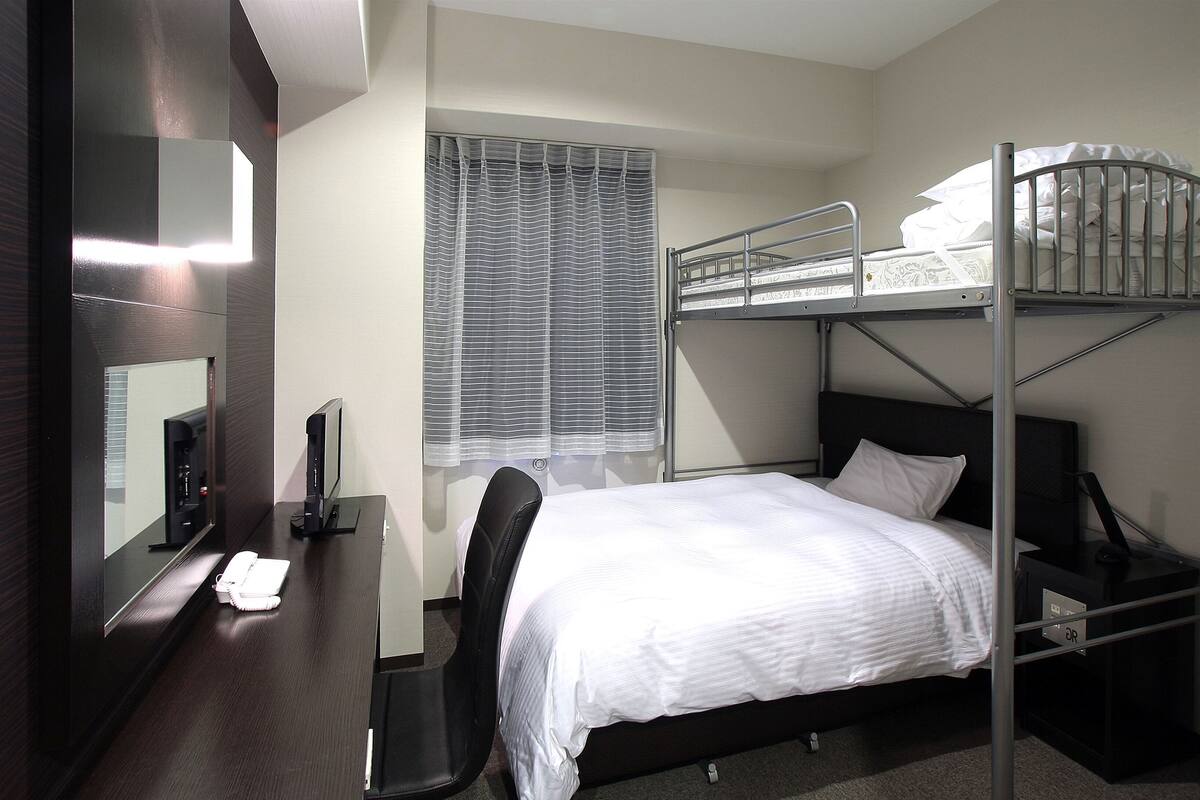 Een slaapkamer met een stapelbed, bureau, stoel en een raam met jaloezieën.
