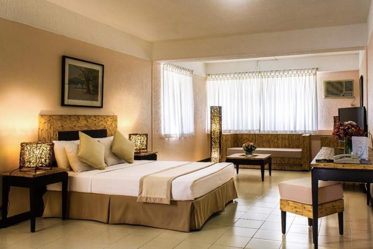 A hotel room with a bed, bedside tables, a desk, a chair, a sofa, and a window with curtains.