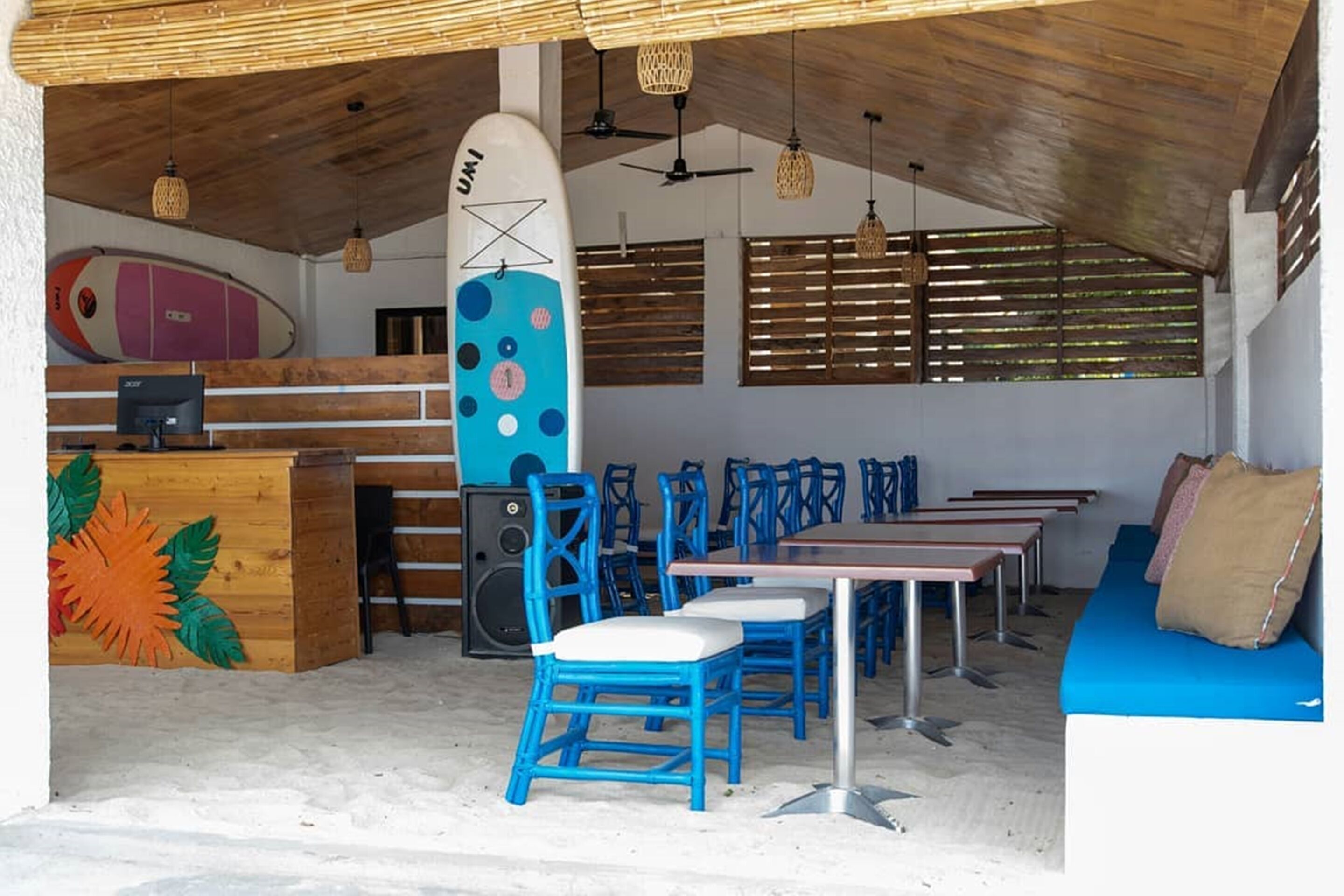 A beachside cafe with wooden tables and blue chairs, a surfboard, and a wooden counter.