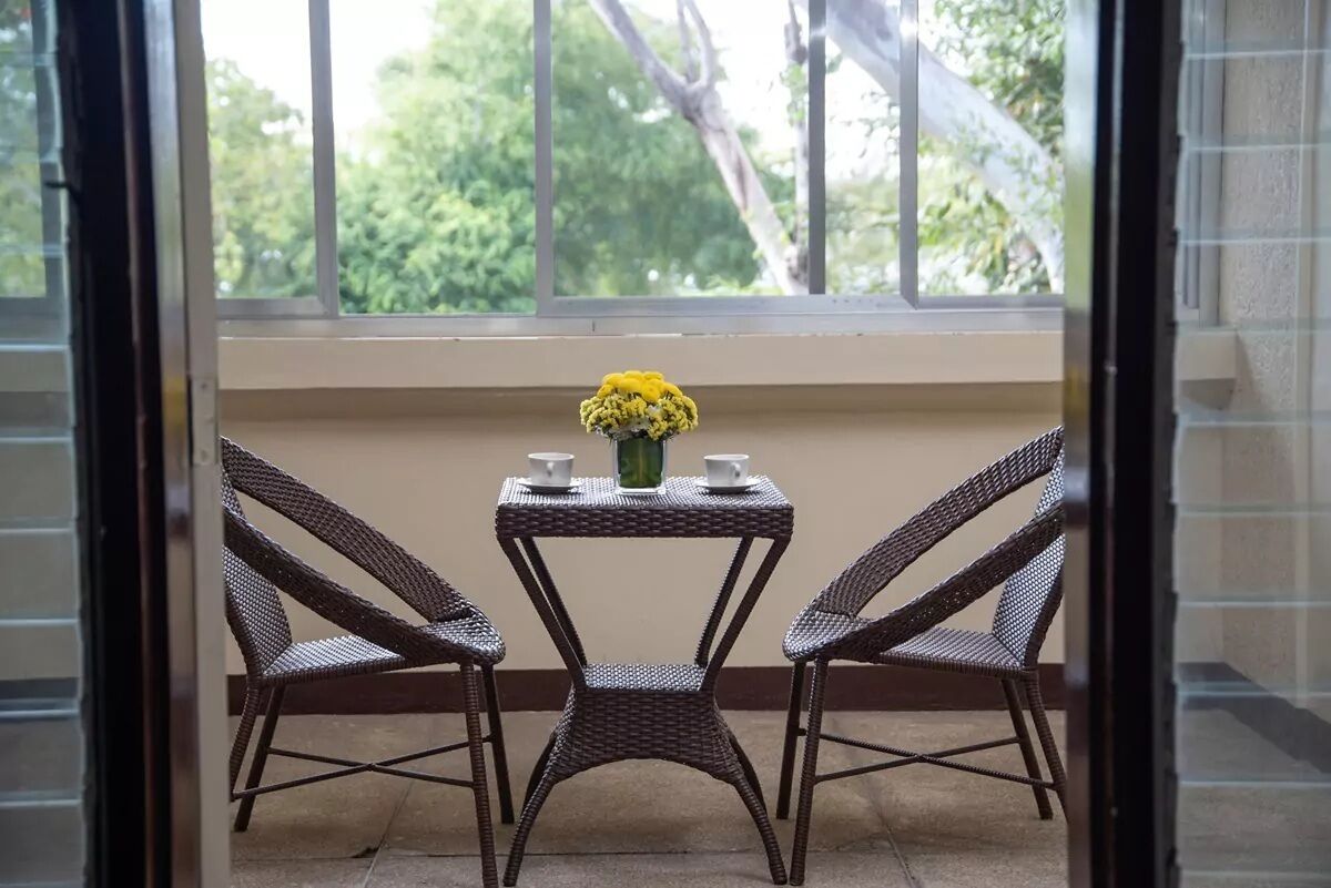 Een klein balkon met rieten stoelen en een tafeltje, een vaas met gele bloemen, en uitzicht op bomen door het raam.