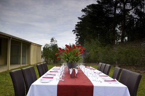 A long dining table set outdoors with a red tablecloth, surrounded by chairs, and a potted plant with red flowers in the center. - Foothills Conference Centre (Mooroolbark)