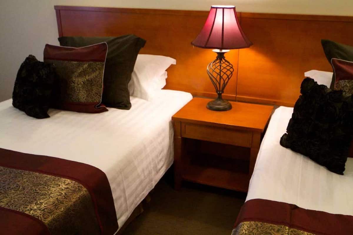 Dos camas individuales con sábanas blancas, cabeceras marrón oscuro y almohadas decorativas. Una mesita de noche de madera con una lámpara de mesa está entre las camas.