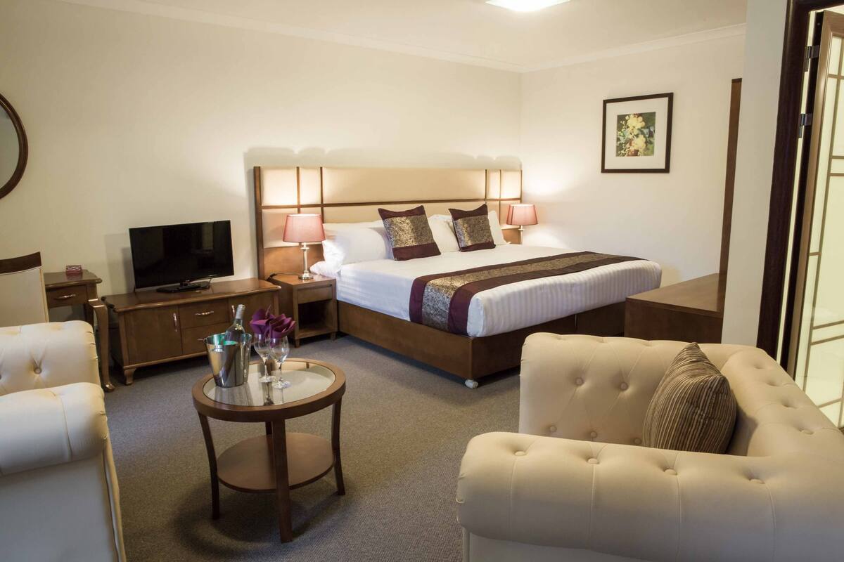 Habitación de hotel con cama, televisor, una mesa redonda con una botella y vasos, y dos sillones.
