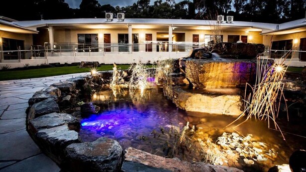 Un edificio moderno con un jardín ajardinado que incluye una fuente de agua y detalles de piedra.