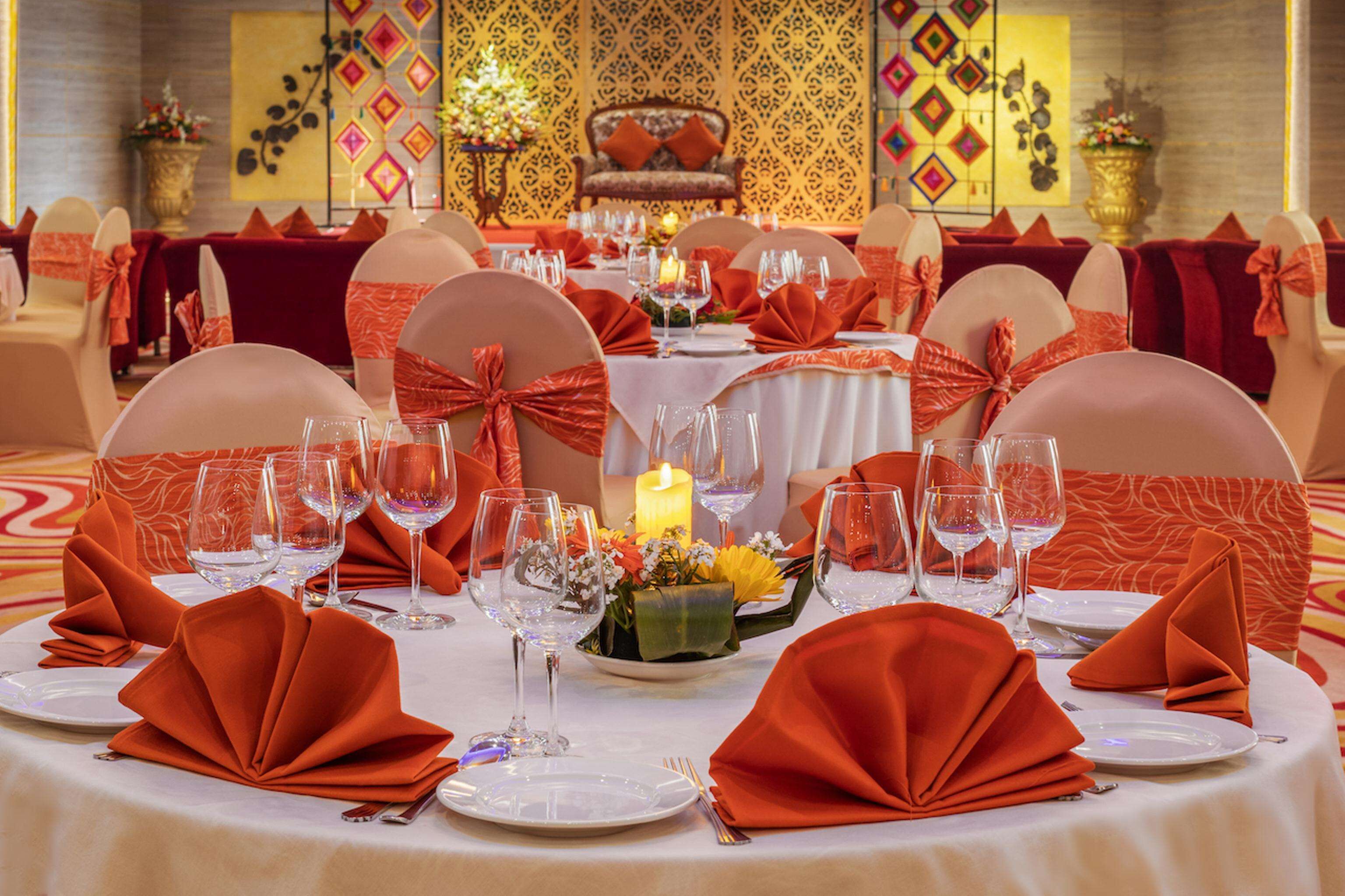 A banquet hall with round tables set for a formal event, featuring white tablecloths, orange napkins, and floral centerpieces.
