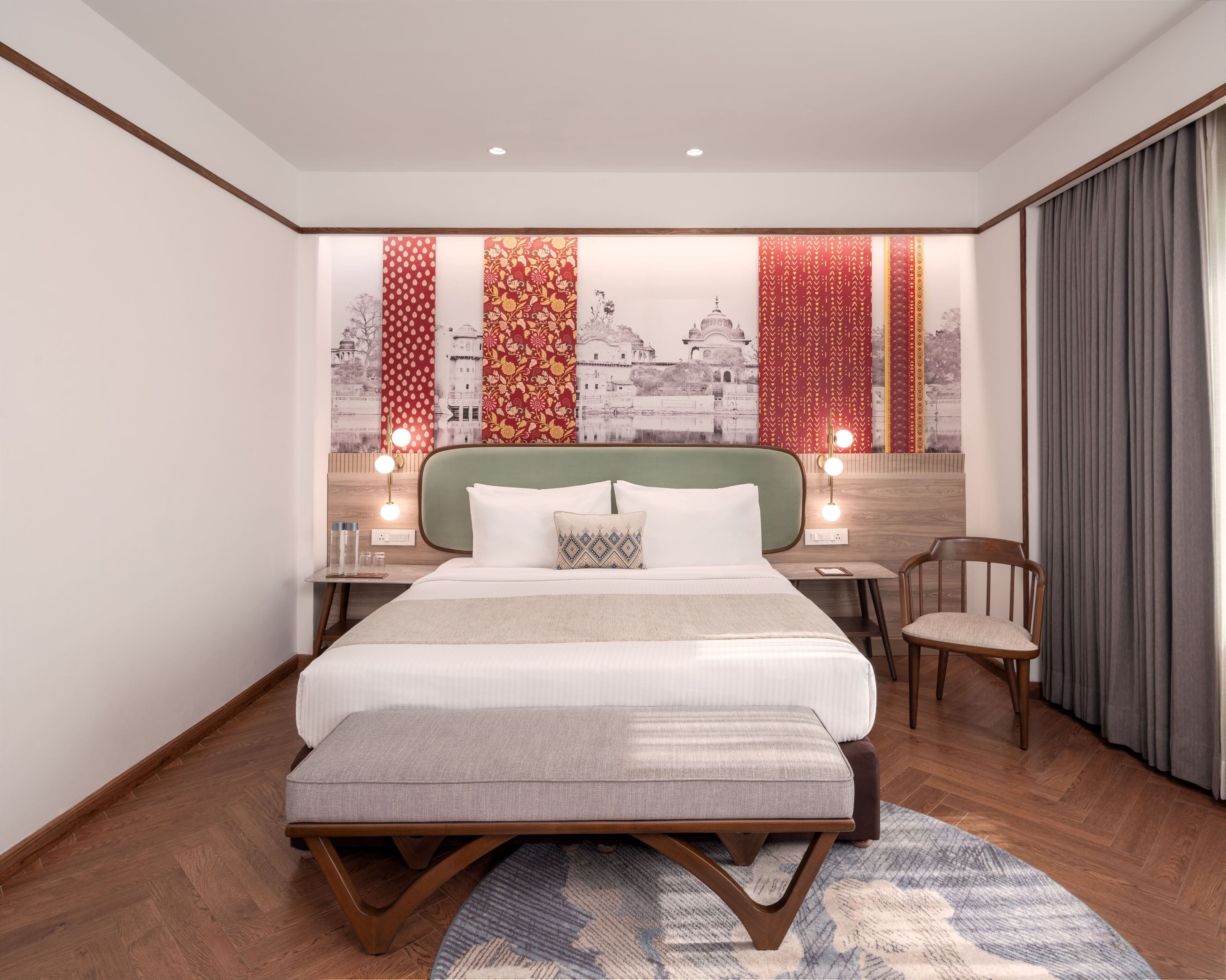 A modern hotel room with a large bed, a wooden headboard, a desk with a chair, and a view of a cityscape through patterned curtains.