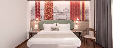 A modern hotel room with a large bed, a wooden headboard, a desk with a chair, and a view of a cityscape through patterned curtains.