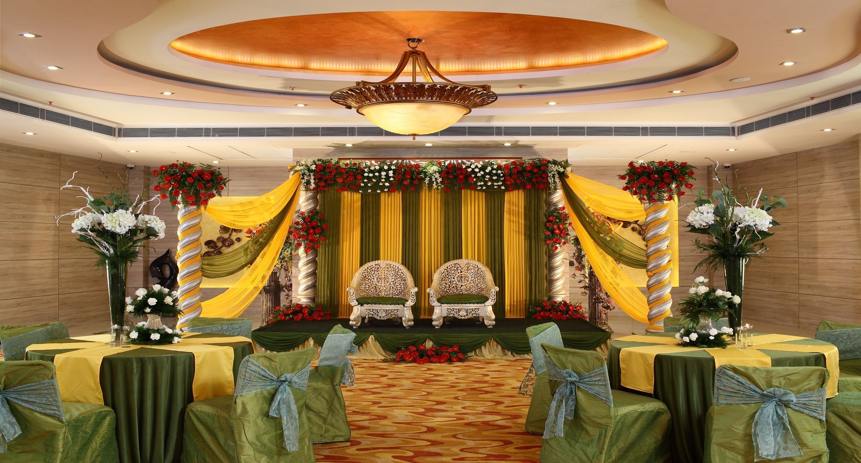 A stage with two ornate chairs, draped with yellow and green fabric, and surrounded by floral arrangements. Tables with green tablecloths and blue sashes are arranged in the foreground.
