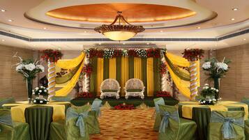 A stage with two ornate chairs, draped with yellow and green fabric, and surrounded by floral arrangements. Tables with green tablecloths and blue sashes are arranged in the foreground.