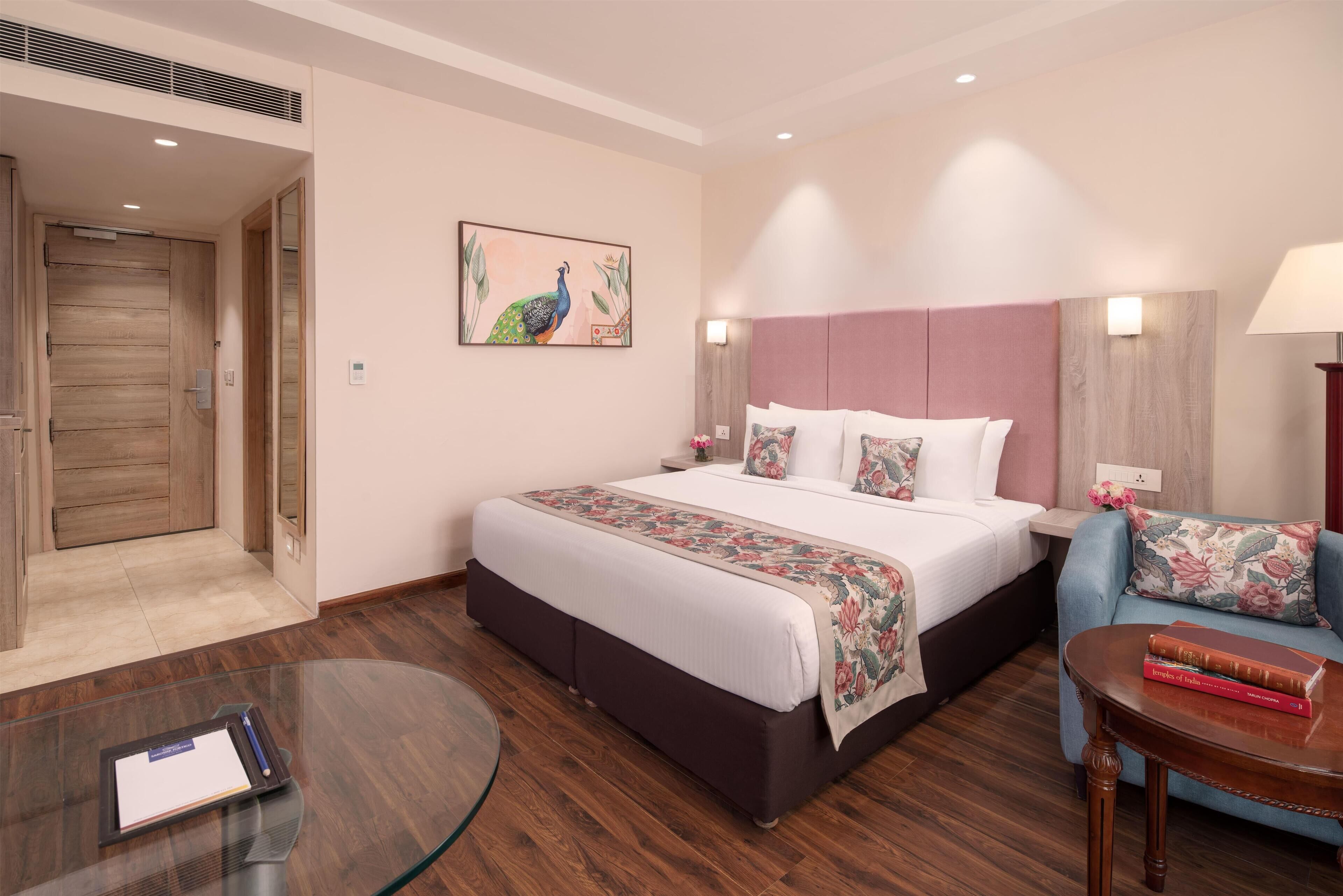 A hotel room with a large bed, a desk with a glass top, a chair, and a small table with books.