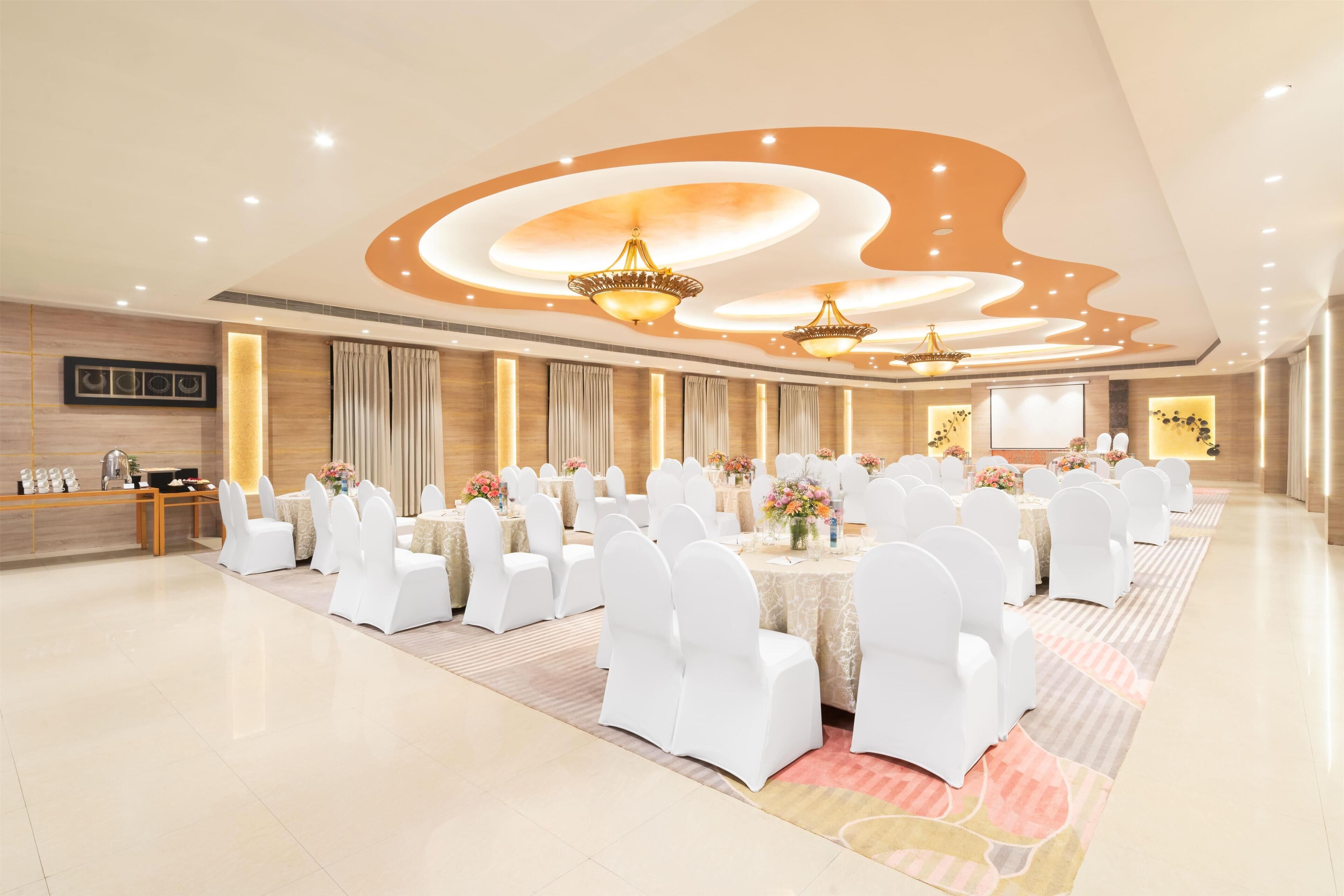 Un amplio salón de banquetes con mesas redondas, sillas blancas y elegantes lámparas de araña.