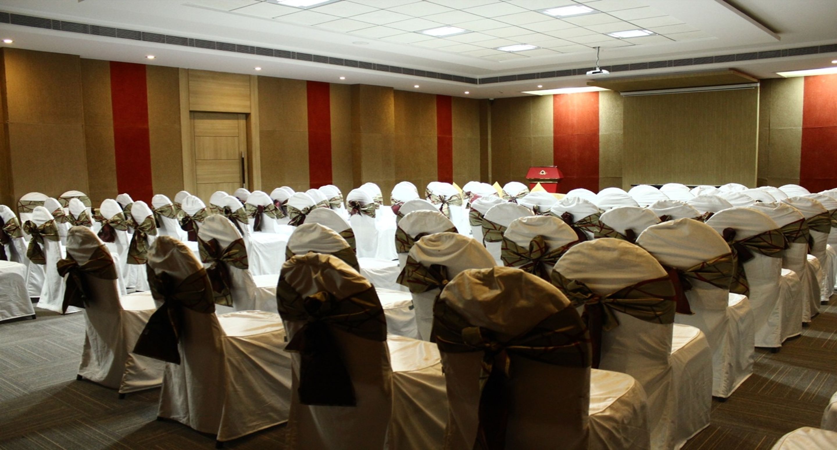 A conference room with rows of tables and chairs, each with white tablecloths and white chairs tied with brown sashes.